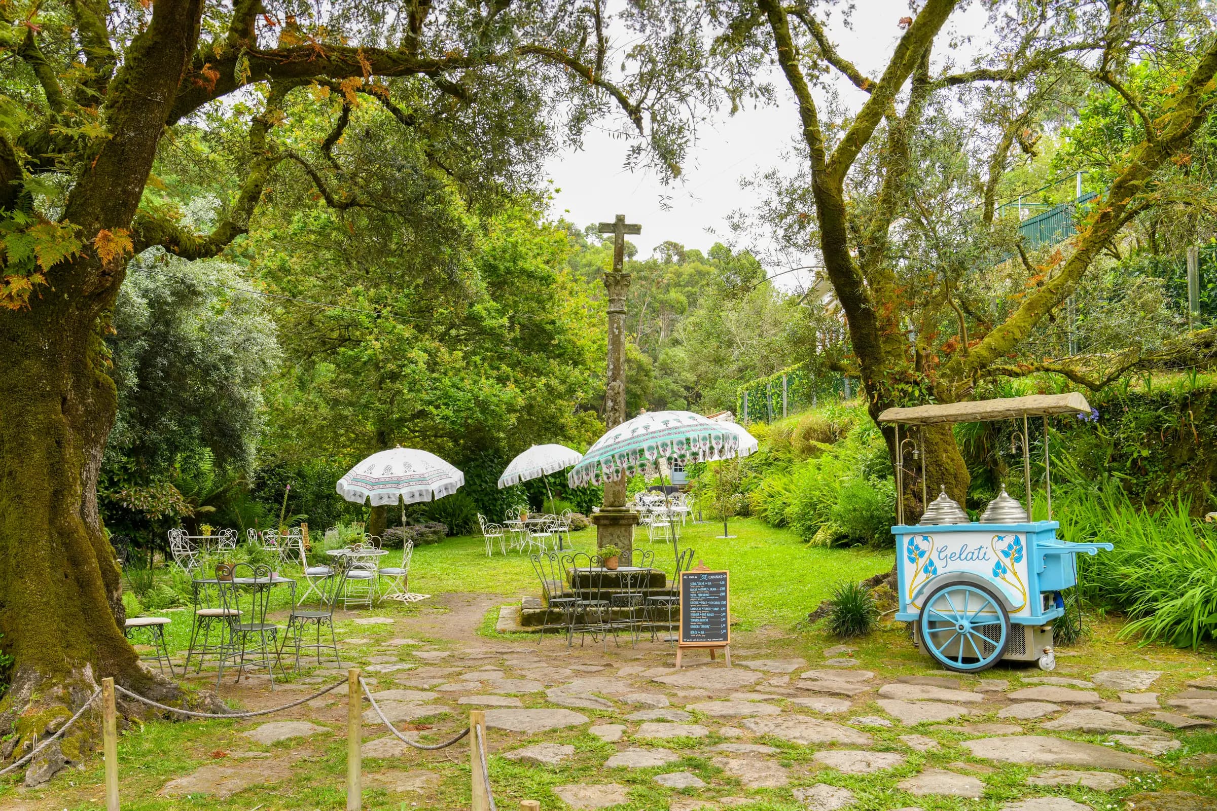 Café tables and white parasols under the trees, by the stone cross